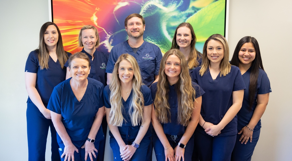 Dental team of nine women in blue scrubs posing together in front of colorful abstract artwork.