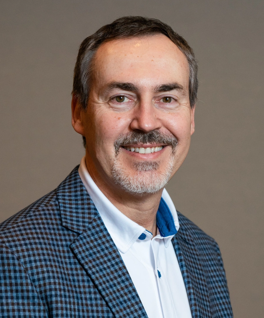 Headshot of a smiling middle-aged man with short brown hair, brown eyes, a mustache, and a grey-and-white goatee. He is wearing a blue-and-white patterned gingham blazer over a white button-down shirt with a blue inner collar. He is positioned from the chest up, angled slightly to the right, against a neutral, solid grey background.